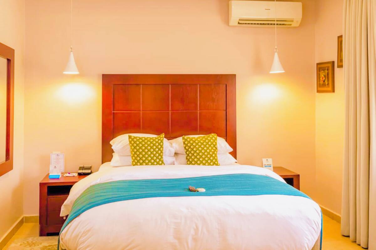 A hotel room with a wooden headboard, two bedside tables, a bed with white bedding and a blue throw, and an air conditioning unit.