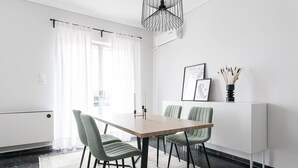 A modern dining area with a wooden table, four upholstered chairs, a black ceiling light, and a white wall with a framed picture and a vase with dried plants. - Spacious 1BD Apartment near HELEXPO Marousi (Marousi)