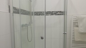 A glass-enclosed shower stall with a tiled floor and a white towel hanging on a radiator. - Trang Hotel (Rastatt)