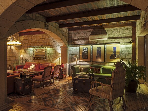 Un salon accueillant avec un mur en pierre, un plafond en bois et une cheminée. On y trouve des sièges confortables, notamment un canapé et des fauteuils, ainsi qu'une petite table avec une lampe.