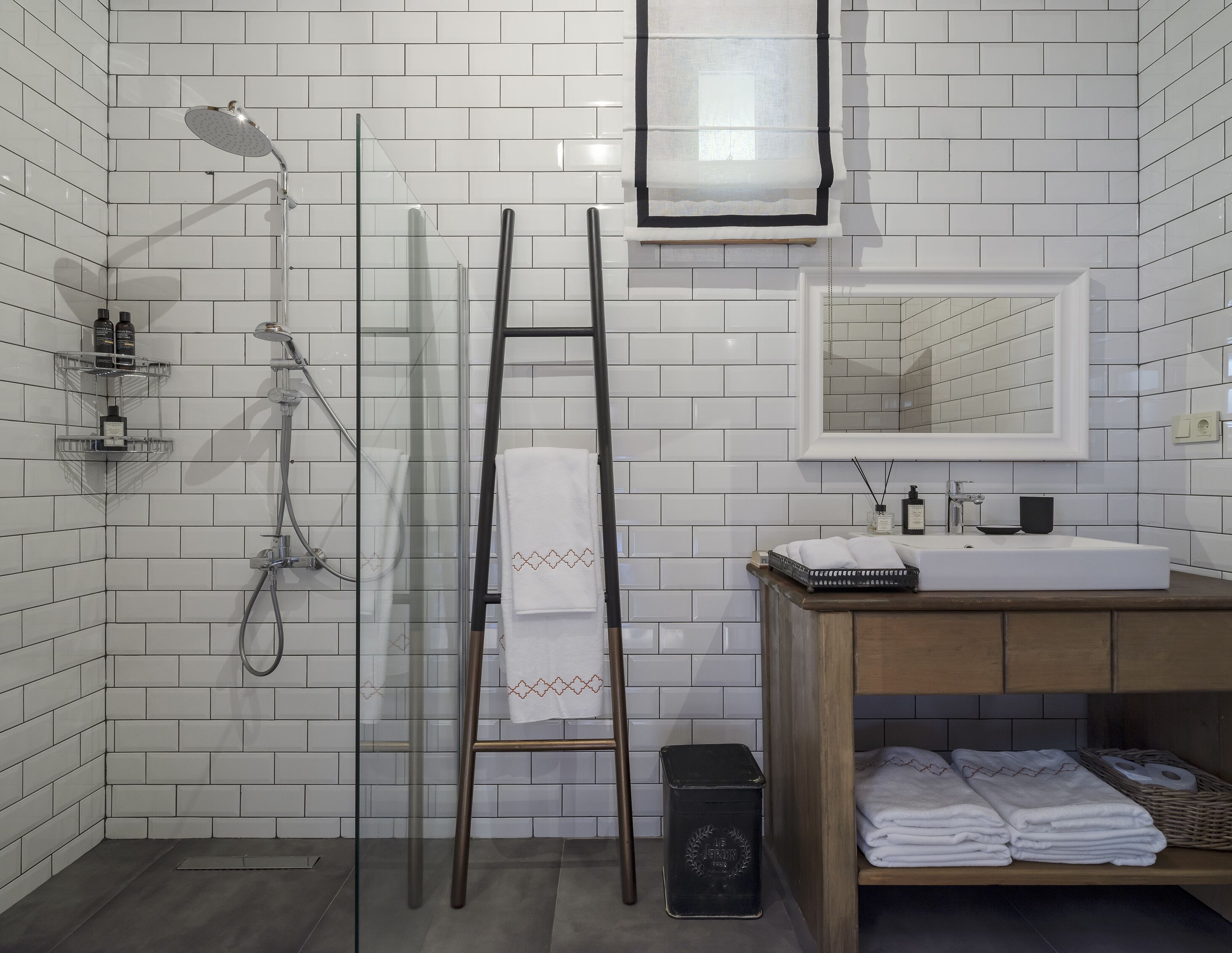 Un baño con paredes de azulejos blancos, cabina de ducha de vidrio, tocador de madera con lavabo y espejo sobre el tocador.
