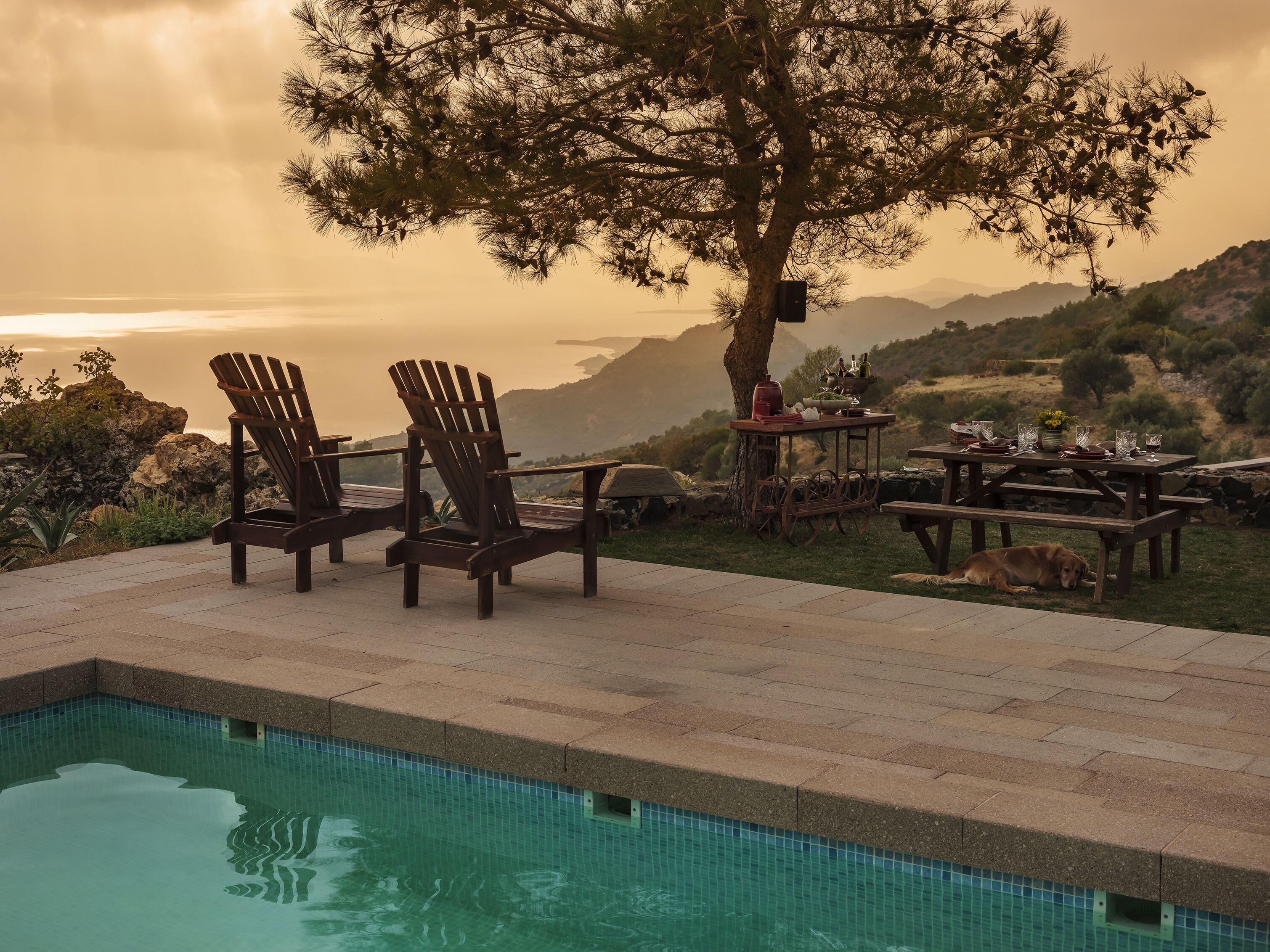 Zona junto a la piscina con dos sillas tipo Adirondack, una mesa de picnic y un perro acostado cerca.