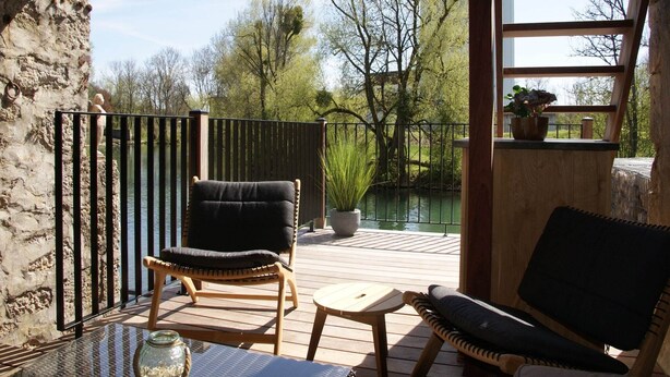 A wooden deck with two wicker chairs and a small table, overlooking a body of water and trees.