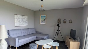 Un salon moderne avec un canapé gris, une petite table ronde, deux tabourets, une carte, un tableau encadré, un trépied, un téléviseur et une lampe.