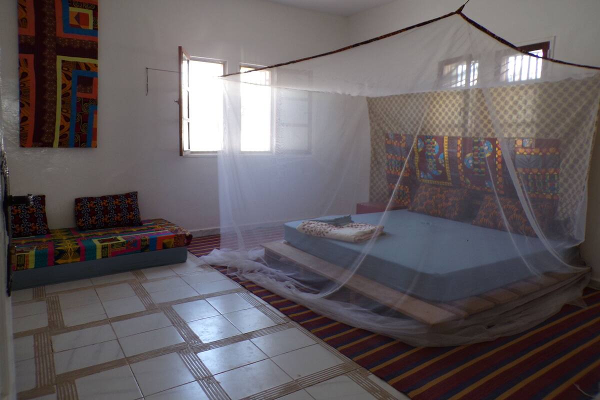 A room with a bed under a mosquito net, a colorful quilt on the wall, and a patterned rug.
