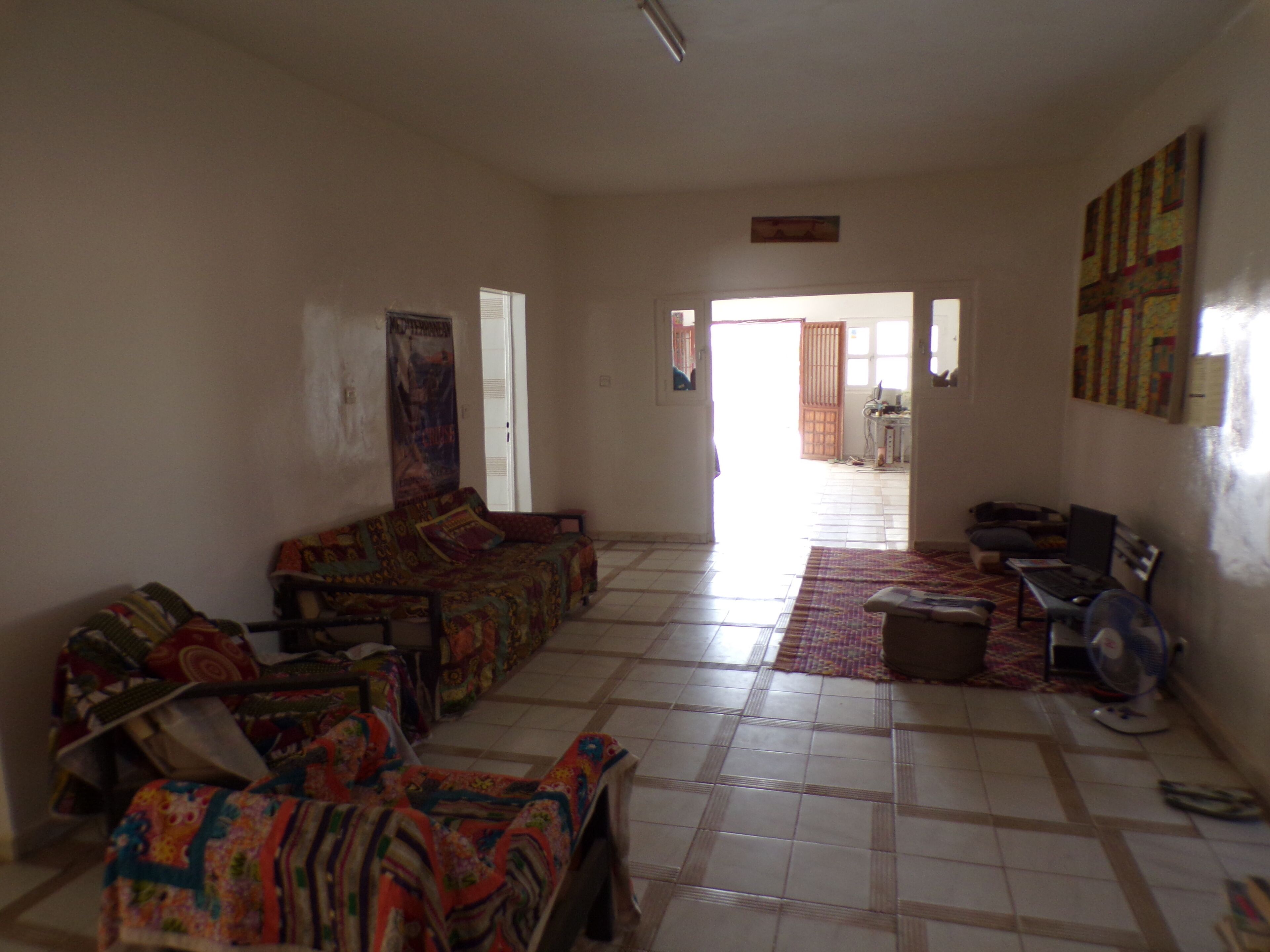 A spacious living room with a sofa set, a small table, and a fan. There is a door leading to another room and a window with blinds.