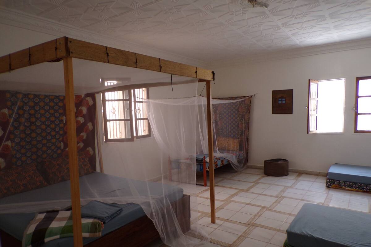 A room with a wooden bed frame, a mosquito net, and a patterned wall. There are two windows and a door.