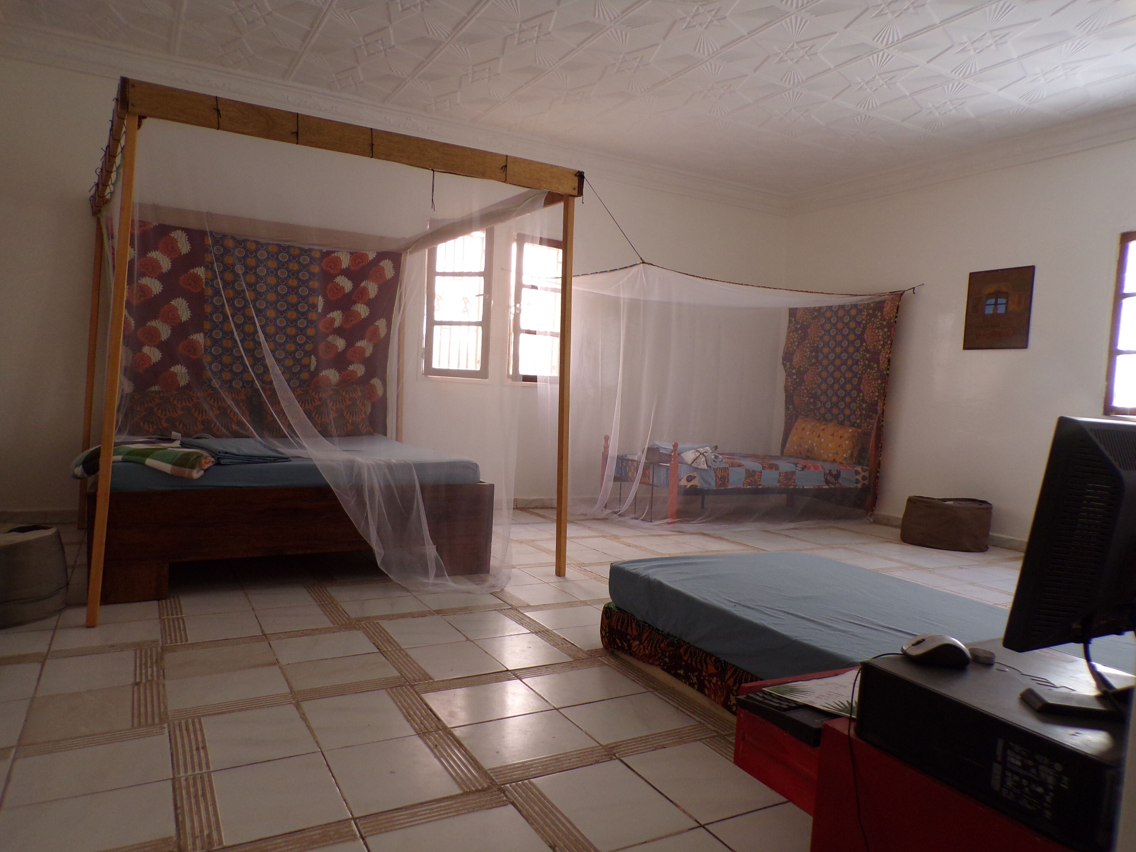 Une chambre avec deux lits, dont un à baldaquin et un autre sans, un bureau avec un ordinateur et un écran, et une poubelle.