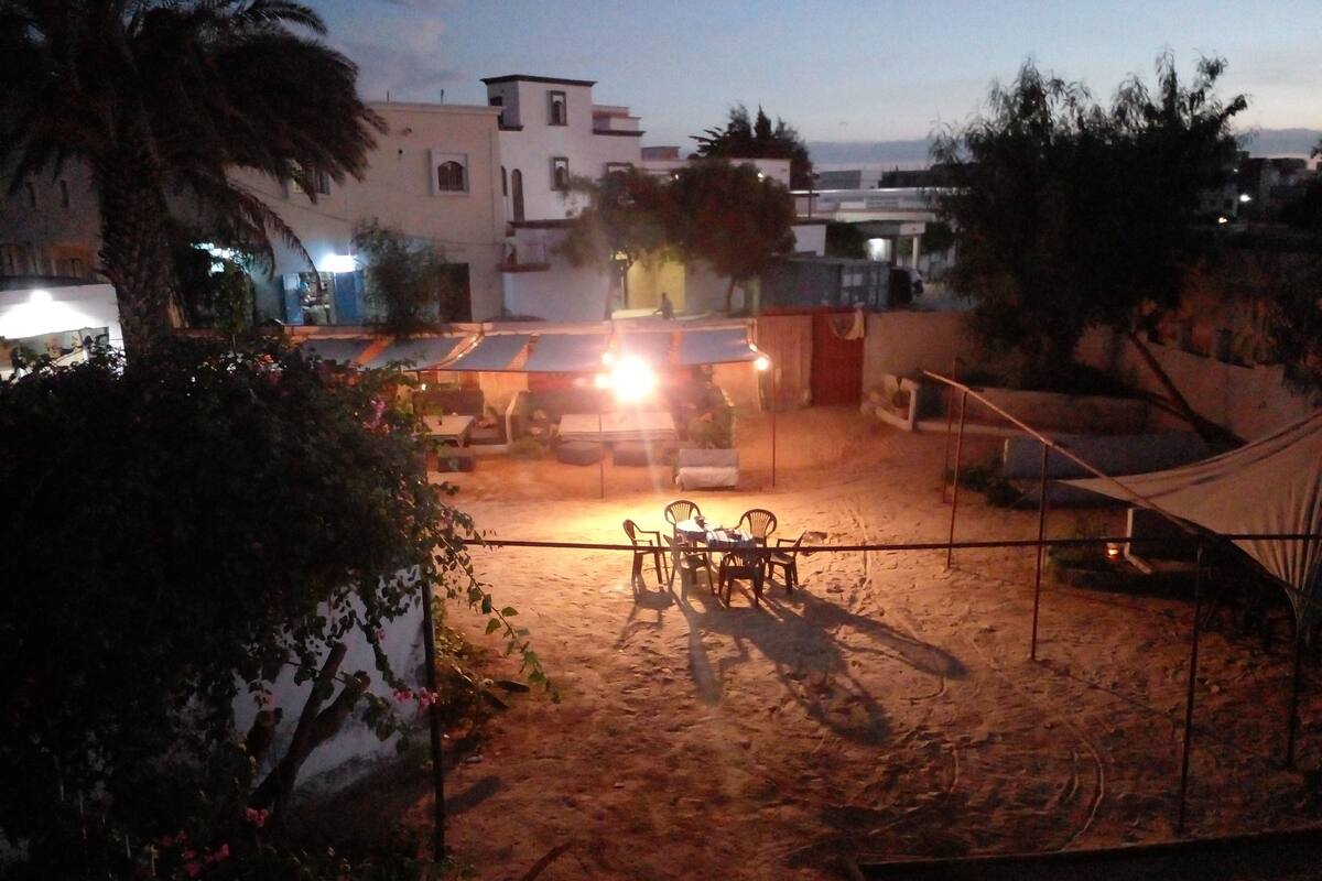 A sandy outdoor area with a table and chairs, surrounded by buildings and palm trees.