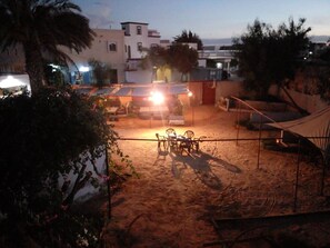 A sandy outdoor area with a table and chairs, surrounded by buildings and palm trees. - Le Triskell Auberge - Hostel (Nouakchott)