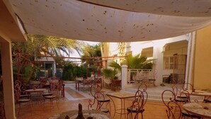 An outdoor patio area with round tables and chairs, surrounded by palm trees and a covered seating area. - Le Triskell Auberge - Hostel (Nouakchott)