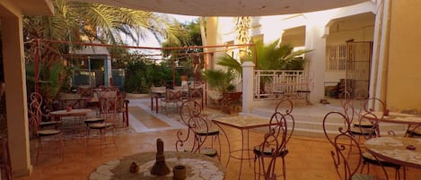 Un patio al aire libre con mesas y sillas redondas, rodeado de palmeras y una zona de estar cubierta.