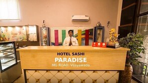Mostrador de recepción en Hotel Sashi Paradise, con vista clara al mostrador, un hombre detrás y una vitrina con artículos.
