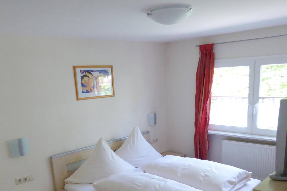 A neatly made bed with white linens in a room with wooden flooring, a small bedside table, a wall-mounted light fixture, and a window with red curtains.