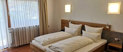 Cama doble con cabecera de madera y ropa de cama blanca en una habitación con suelo de madera, una mesita de noche pequeña y una ventana con cortinas traslúcidas.