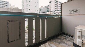 A narrow balcony with a glass barrier, a small air conditioning unit, and a view of surrounding buildings. - Diamante Yakuin (Fukuoka)