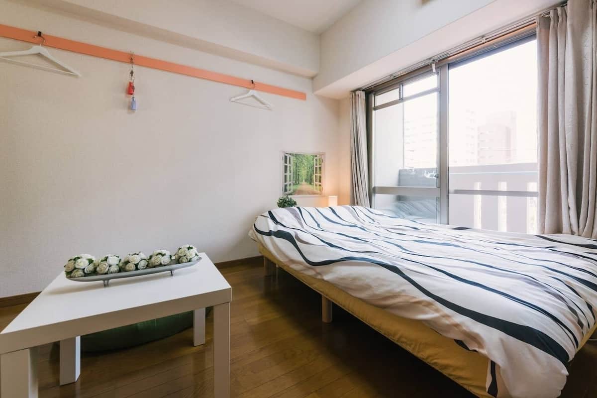Un dormitorio con cama, una mesita con un arreglo floral, una ventana con cortinas y vistas al exterior.