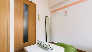 A room with a wooden door, a white table with a flower arrangement, a green cushion, a mirror, and a clothes hanger with items. - Diamante Yakuin (Fukuoka)