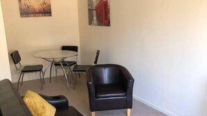 Un salon avec un canapé en cuir noir, deux coussins jaunes, un fauteuil noir, une table basse en verre et un tableau représentant une cabine téléphonique rouge accroché au mur.