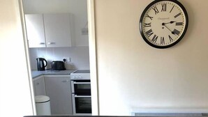 Un canapé en cuir noir agrémenté de deux coussins jaunes, une horloge murale et une vue sur un coin cuisine avec des meubles et des appareils électroménagers blancs.