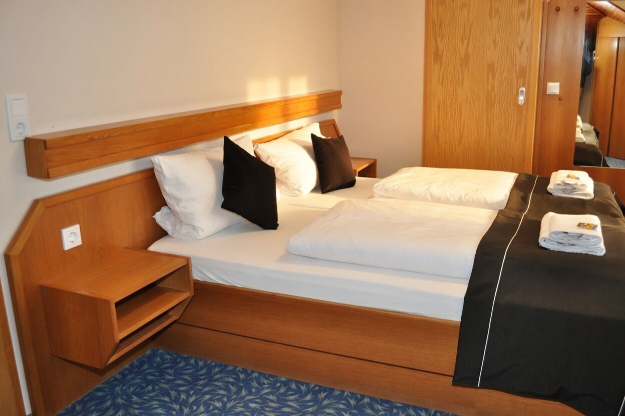 A hotel room with a wooden headboard, a bed with white bedding, a blue patterned rug, and a wooden nightstand.