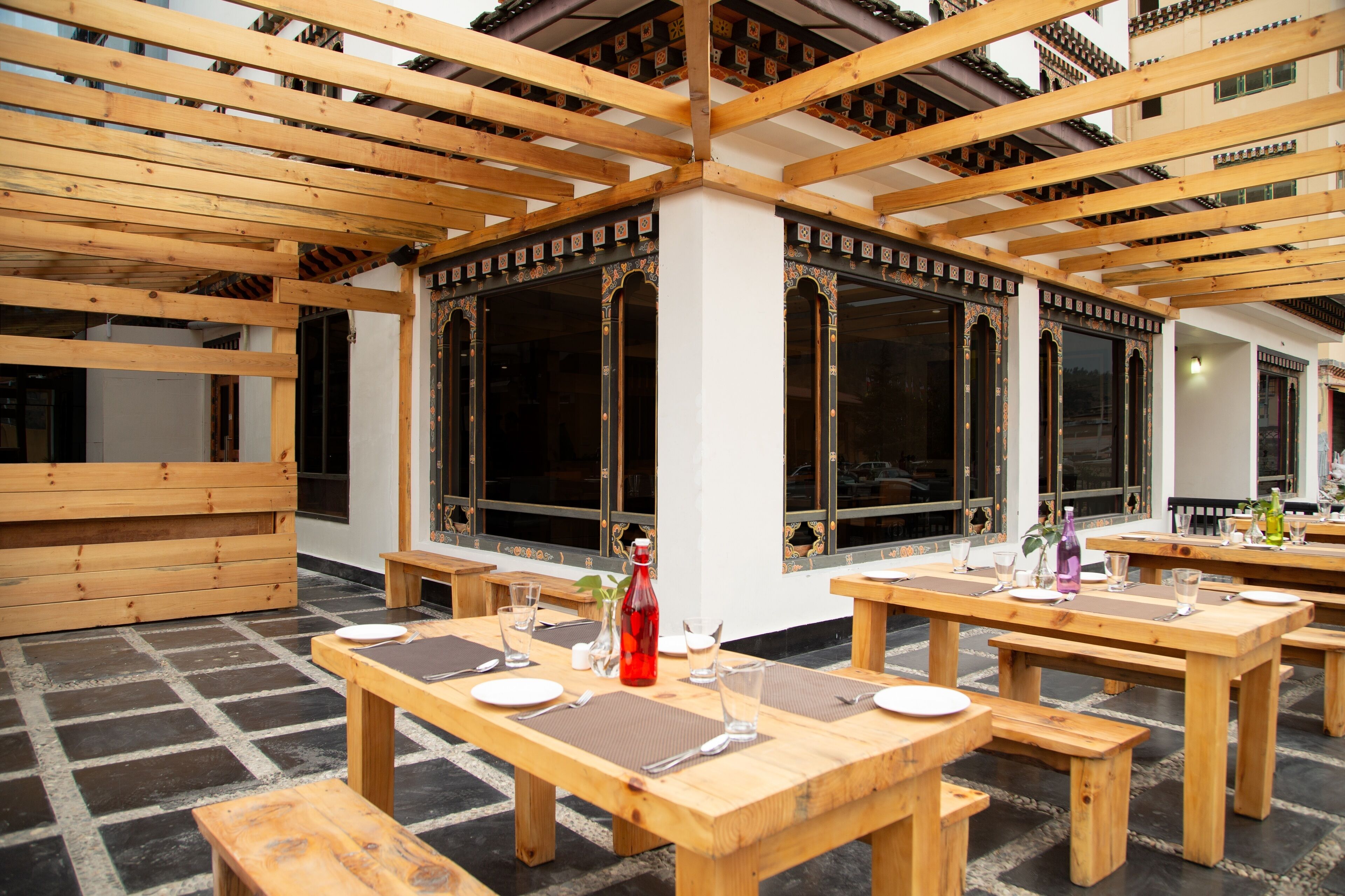 An outdoor dining area with wooden tables and benches, large windows, and a traditional architectural style.