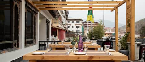 An outdoor dining area with wooden tables and benches, a flag, and buildings in the background.