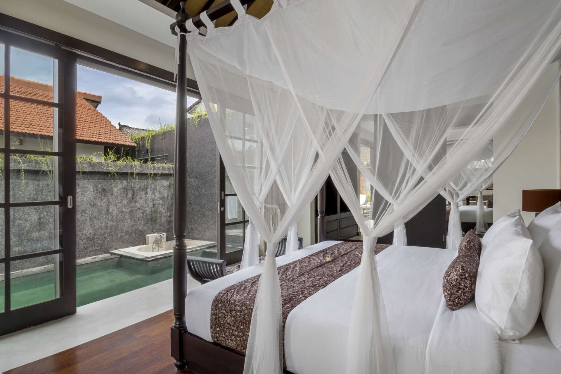 A bedroom with a four-poster bed, a glass door leading to a pool, and a view of a building with a tiled roof.