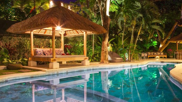 Un espace aménagé au bord de la piscine, avec un gazebo à toit de chaume, des chaises longues et une piscine qui reflète la verdure environnante.