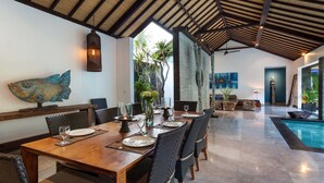 A dining area with a wooden table set for four, surrounded by wicker chairs. There is a large fish decoration on the wall, a hanging light fixture, and a view of a swimming pool in the background. - Bali Villa Near the Beach, 2080 (Seminyak)