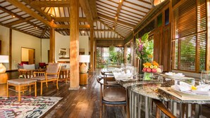 A spacious dining area with wooden beams, a long table set for dining, and a view of greenery outside.