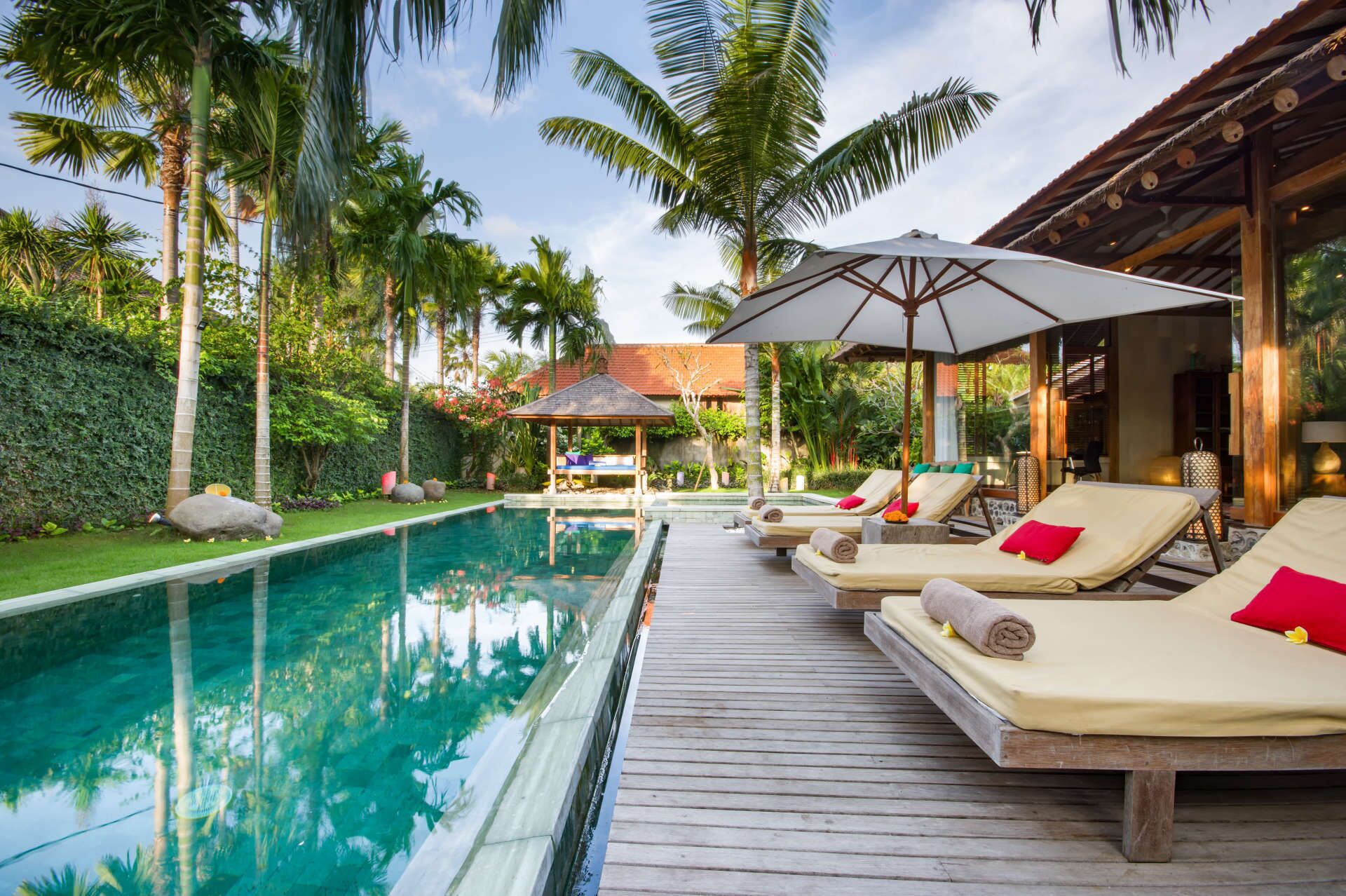 Zona junto a la piscina con sillones de descanso y sombrillas, rodeada de palmeras y una terraza de madera.