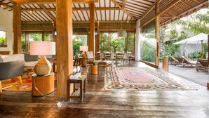 A spacious indoor area with wooden beams, a dining setup, and a view of lush greenery outside.