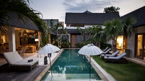 A pool area with lounge chairs and umbrellas, surrounded by palm trees and a building with a tiled roof.