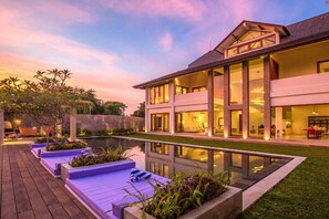 A modern two-story house with a large glass facade, a swimming pool, and a wooden deck with purple seating.