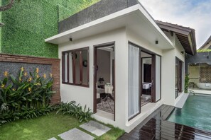 Ein modernes Haus mit Swimmingpool, mit Glastür, einer kleinen Sitzecke mit Tisch und Stühlen sowie einem Garten mit tropischen Pflanzen.
