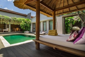 Zona junto a la piscina con un muelle de madera, una estructura de techo de paja y una silla de descanso con cojines.