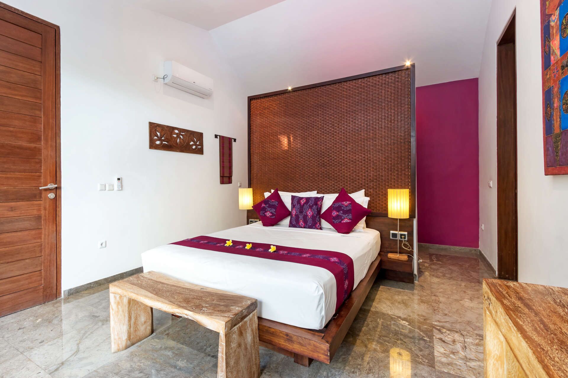 A hotel room with a wooden bed, a bench, a wall-mounted air conditioner, and a decorative panel.
