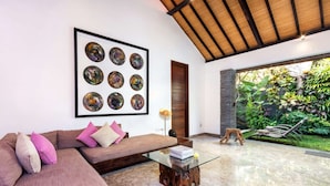 A modern living room with a large framed abstract art piece, a wooden ceiling, a glass coffee table, and a view of a lush garden.