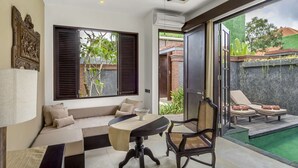 A modern room with a sofa, a dining table, and a chair. There is a view of a swimming pool and a garden outside. - Villa for Rent in Bali 2010 (Kerobokan)