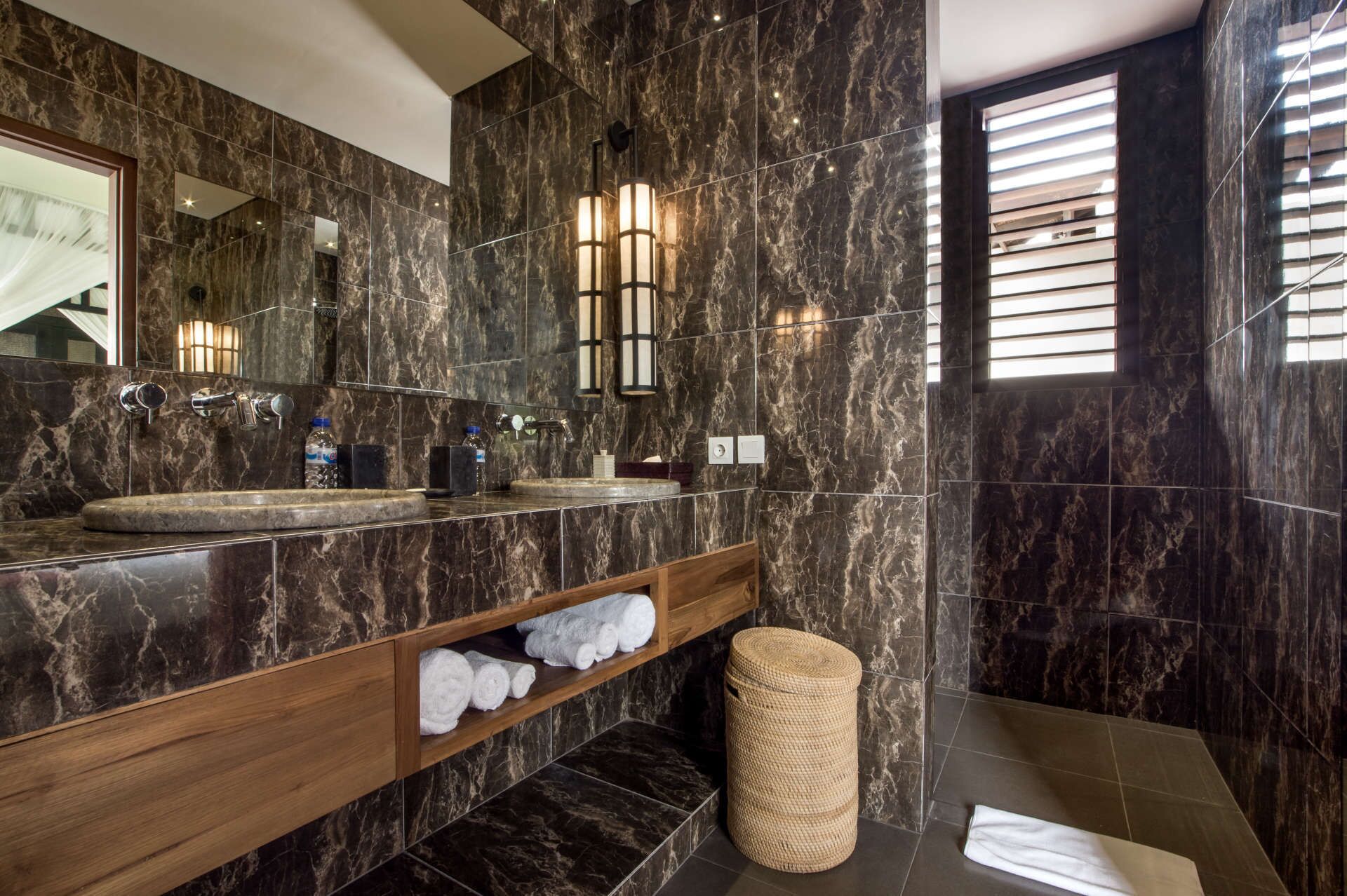 A modern bathroom with dark marble tiles, two sinks, a wooden vanity, and a large mirror.