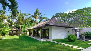A single-story building with a thatched roof, surrounded by lush greenery and palm trees.