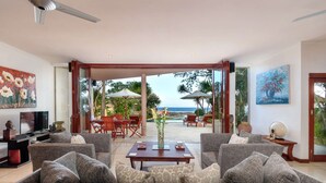 A spacious living room with a large sofa, a coffee table, and a TV. There is a dining area with chairs and a table, and a view of the ocean through large windows. - Villa for Rent in Bali 2002 (Manggis)