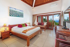 A spacious bedroom with a large bed, a wooden desk, and a balcony with a view of the ocean. - Villa for Rent in Bali 2002 (Manggis)