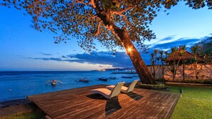 A wooden deck with lounge chairs overlooking a calm sea with boats, a tree with a glowing trunk, and a coastal building in the background. - Villa for Rent in Bali 2002 (Manggis)