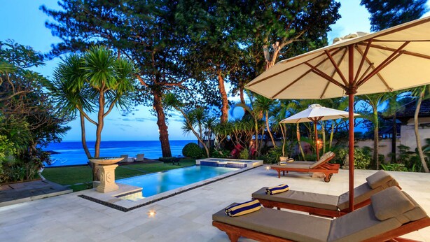 A poolside area with lounge chairs and an umbrella, surrounded by palm trees and a view of the ocean. - Villa for Rent in Bali 2002 (Manggis)