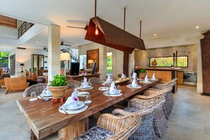 A spacious dining area with a long wooden table set for a meal, surrounded by wicker chairs. A modern kitchen with a large island and stainless steel appliances is visible in the background.