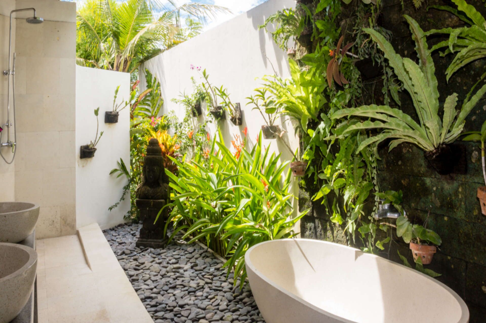 A bathroom with a large bathtub, a shower, and a small sink. There are various plants and a stone wall.