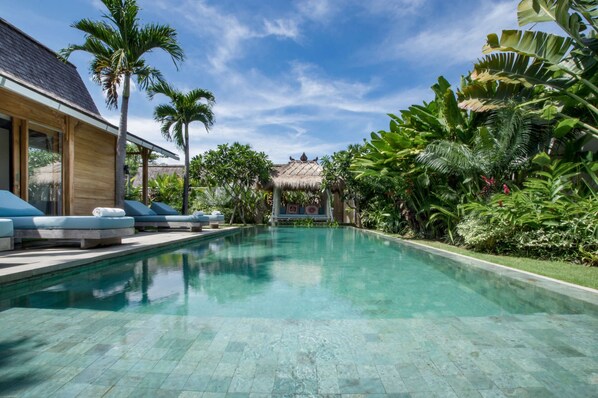 Une piscine entourée de chaises longues, bordée d’une végétation tropicale luxuriante et surmontée d’une structure traditionnelle à toit de chaume.