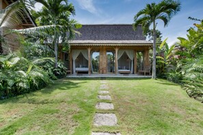Un complexe hôtelier tropical doté d’un espace extérieur couvert avec des sièges, d’un chemin de pierre menant à l’entrée et d’une végétation luxuriante qui entoure l’établissement.